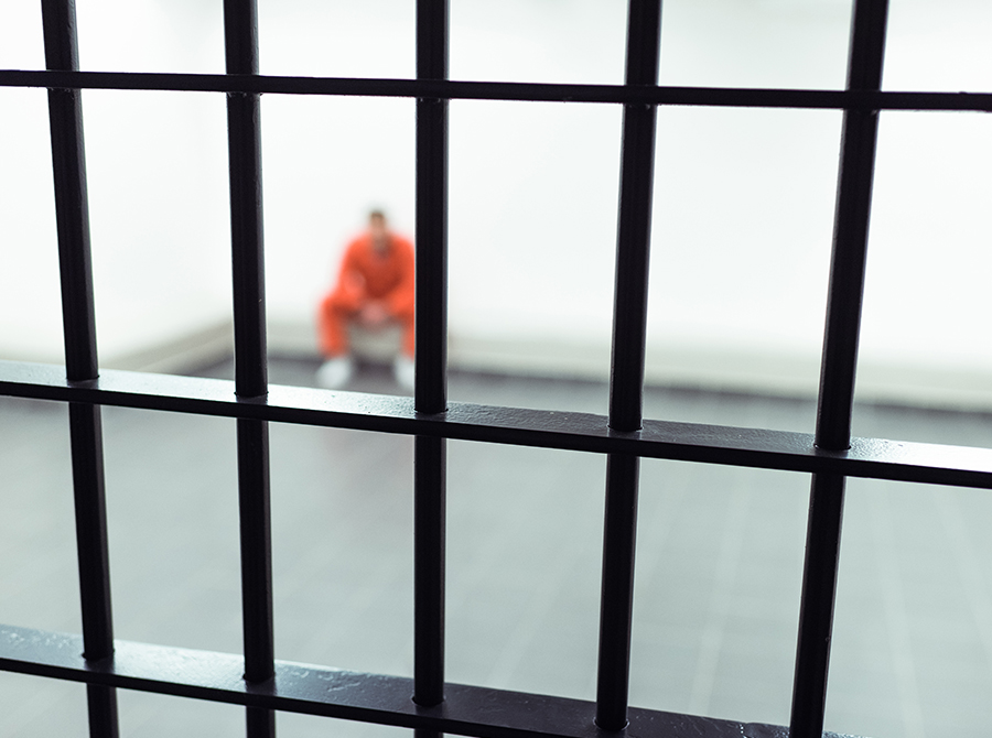 A person in an orange jumpsuit sits behind metal bars in a jail cell, illustrating the line between firm and abusive in corrections through inmate restraint and confinement.
